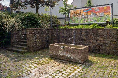 Bild zeigt Ausschnitt vom Dorfplatz von Bitburg Mötsch mit Brunnen im Vordergrund